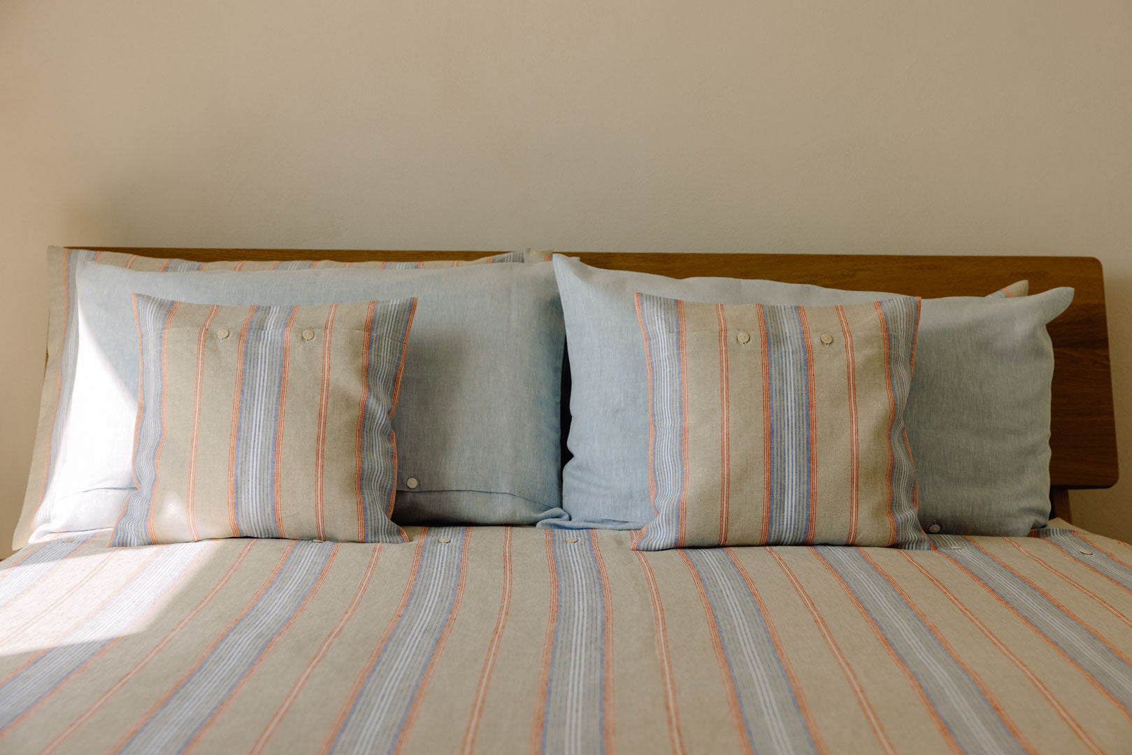 Doppelbett mit Holzkopfteil, darauf vier Kissen und eine Doppeldecke bezogen mit gestreifter und unifarbener Leinenbettwäsche