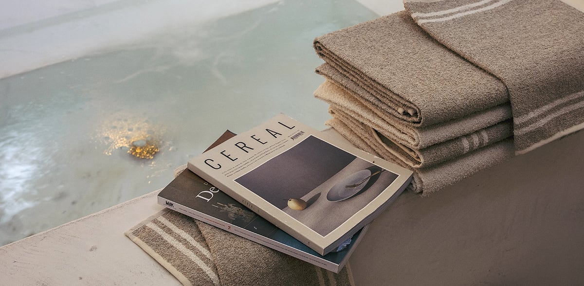 Drei gefaltete beigefarbene Handtücher und ein passendes Handtuch liegen auf dem Rand einer Badewanne. Oben auf den Handtüchern liegen drei Zeitschriften, darunter eine mit Cereal auf dem Cover, neben dem Wasser.