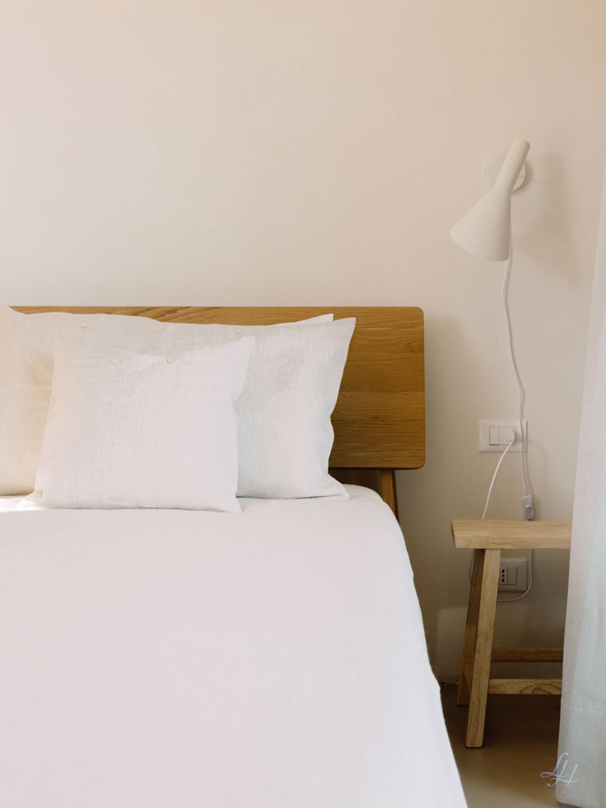 Ein Bett mit weißen Halbleinenkissen und Kissen neben einem hölzernen Nachttisch, unter einer Wandlampe, vor einer beigen Wand.