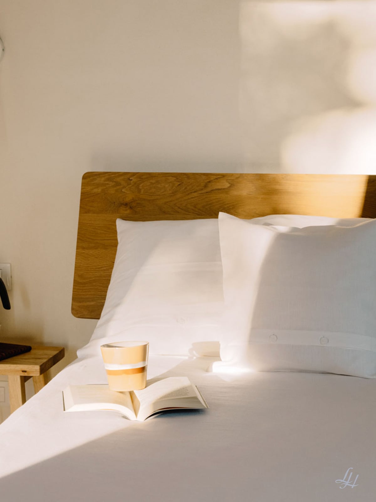 Eine Keramiktasse und ein aufgeschlagenes Buch liegen auf einem Bett mit Halbleinen-Kissen, weißer Bettwäsche und einem hölzernen Kopfteil in sanftem Tageslicht.