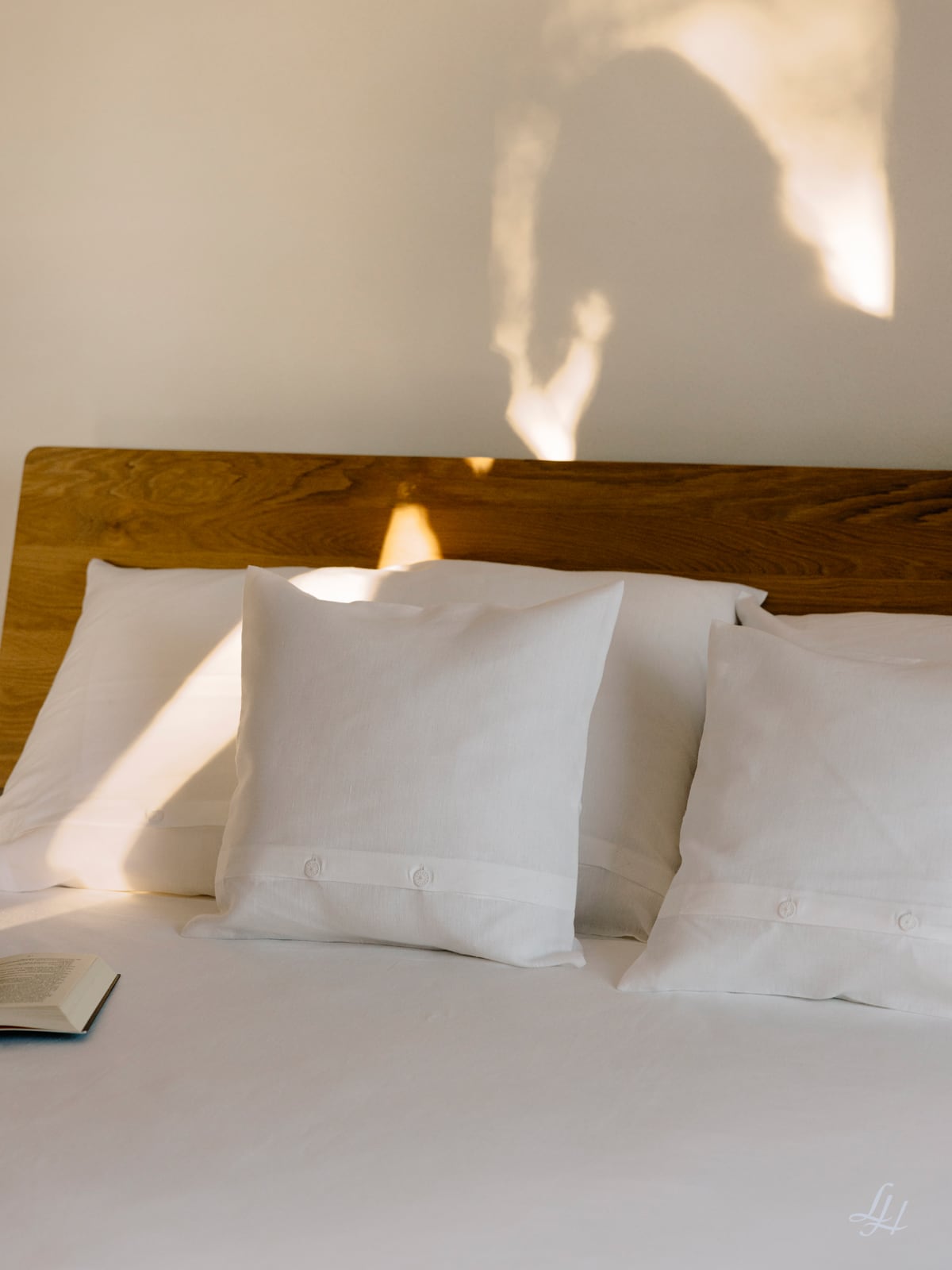 Ein ordentlich gemachtes Bett mit halbleinenen Kissen, weißen Laken, einem hölzernen Kopfteil und einem aufgeschlagenen Buch, das auf dem Bett liegt.