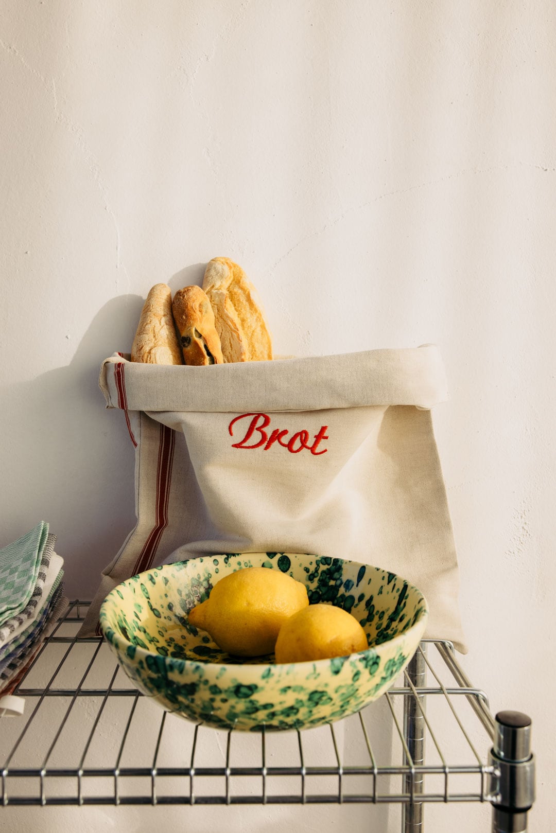 Ein Brotbeutel Halbleinen für natürliche Brotaufbewahrung hält Baguettes; daneben eine gelb-grüne Zitronen-Schale auf dem Metallregal.