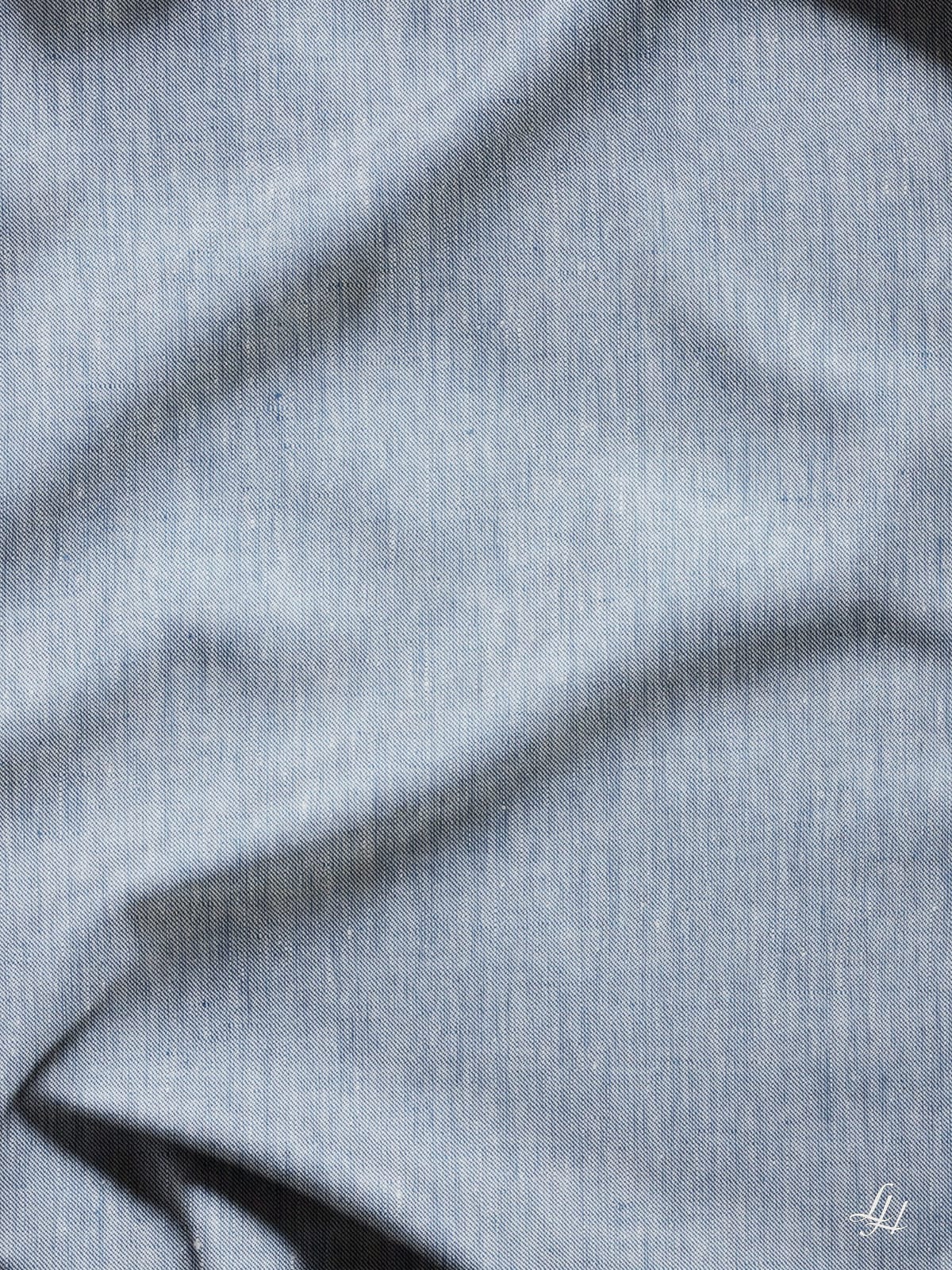 Hellblauer Denim-Stoff mit Köperbindung, weichen Falten und einem kleinen weißen Logo in der rechten unteren Ecke.