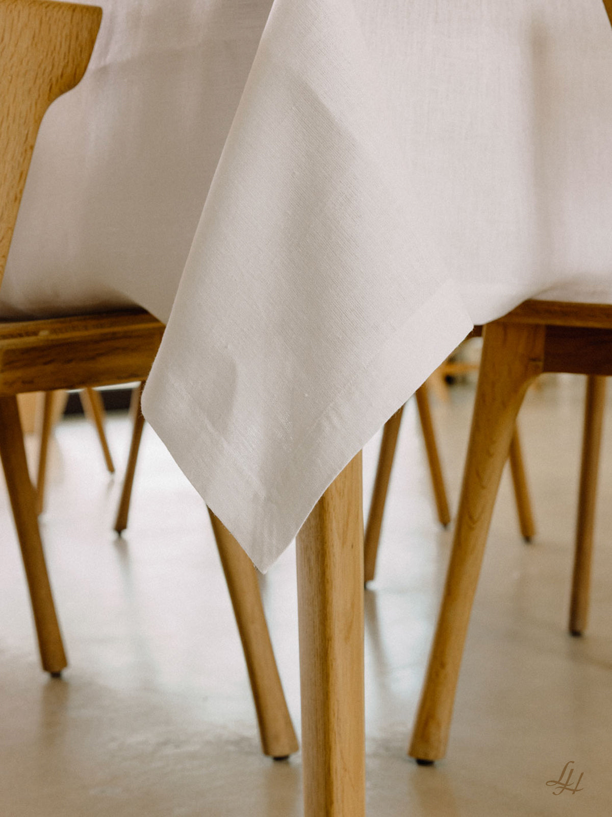 Eine weiße Leinenmanufaktur-Tischdecke liegt über einem Holztisch, umgeben von hellen Stühlen auf einem beigen Boden.