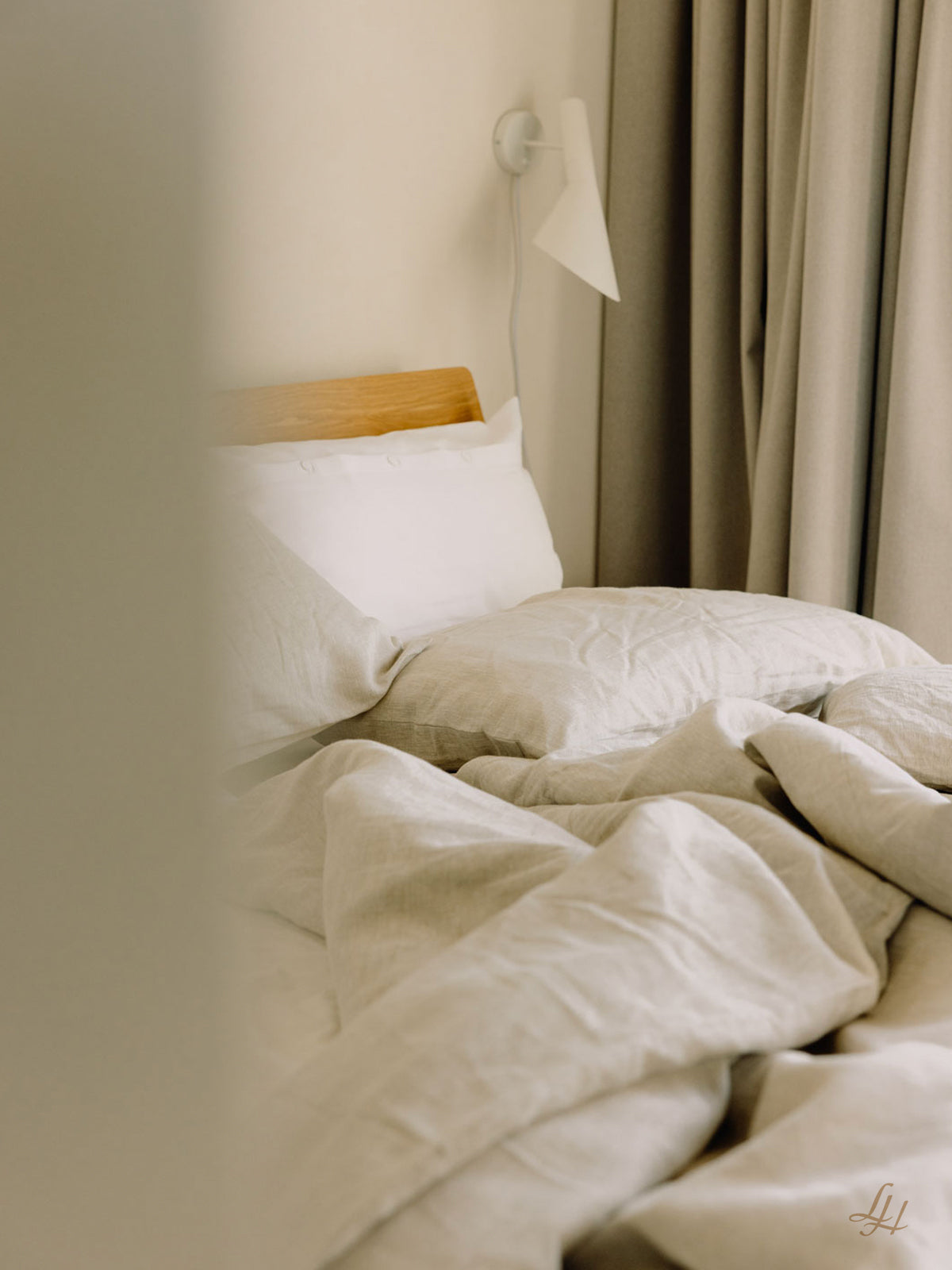 Ein ungemachtes Bett mit beiger Leinenbettwäsche, einem hölzernen Kopfteil, weißen Kissen, einer Wandlampe und beigen Vorhängen im Hintergrund.