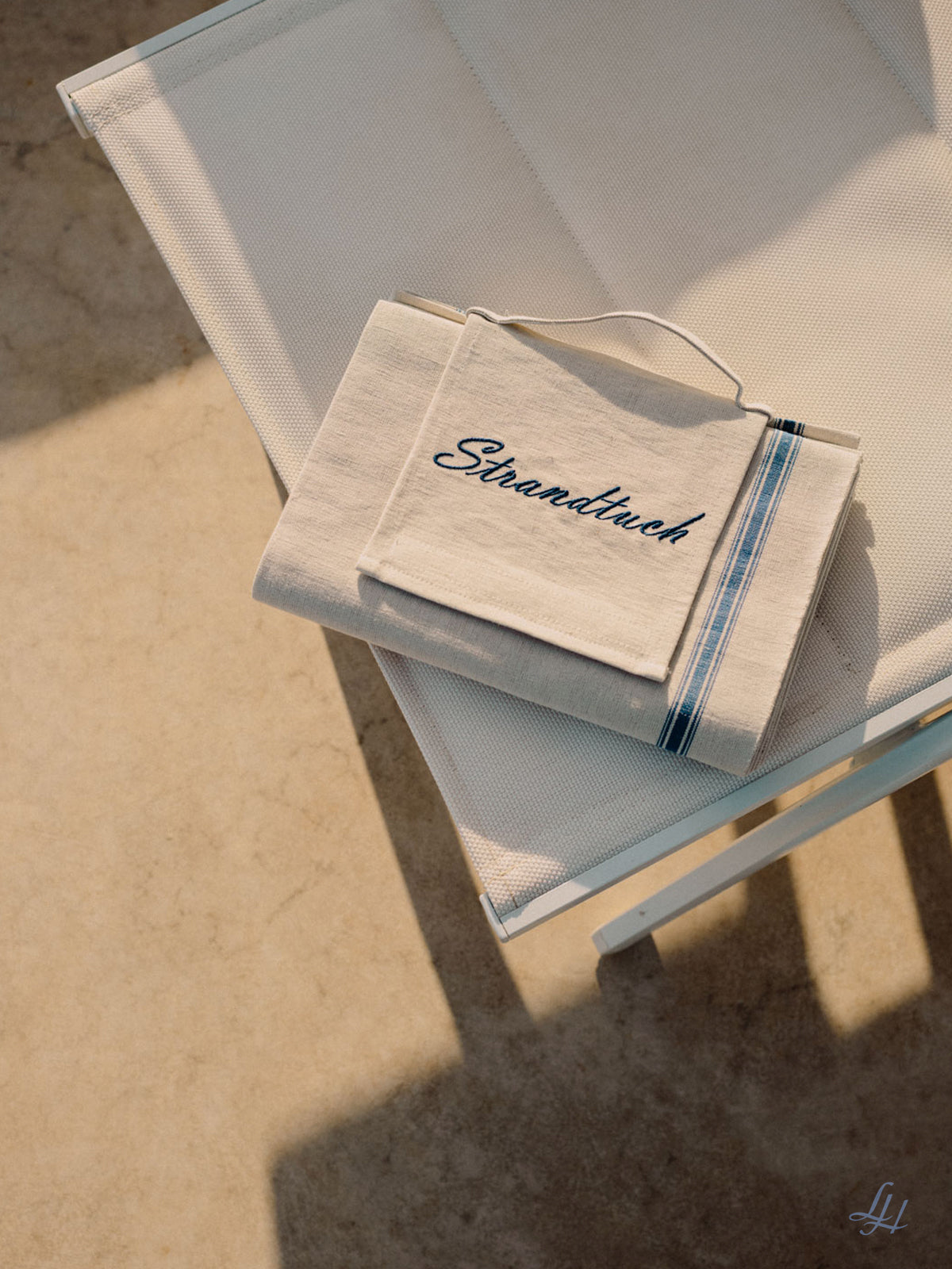 Ein gefaltetes beigefarbenes Halbleinen-Strandtuch mit blauen Streifen und dem aufgestickten Wort "Strandtuch" liegt auf einem weißen Netz-Loungesessel und wirft einen Schatten auf eine Steinfläche.