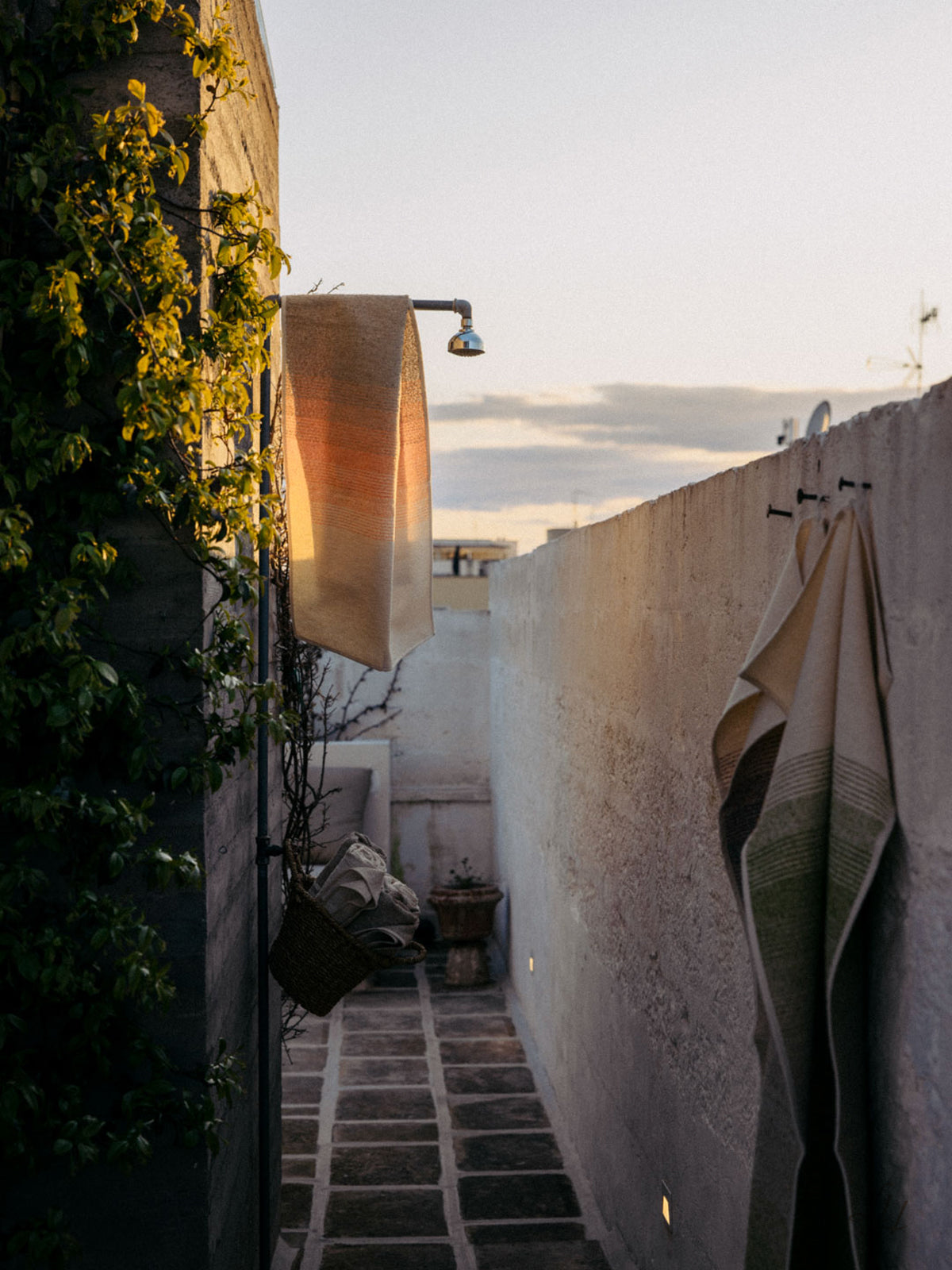 Ein Leinenhandtuch hängt von einer Außendusche an einer Betonwand, das Abendlicht wirft Schatten auf den schmalen Gehweg.