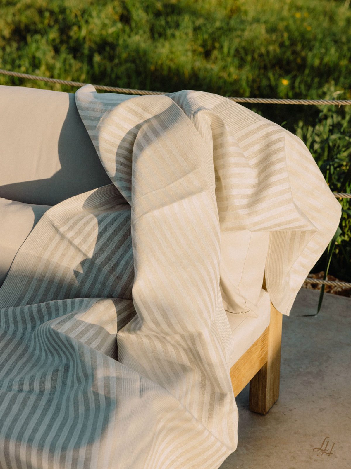 Eine creme- und beigefarbene gestreifte Decke aus europäischem Leinen, drapiert über einem hellen Outdoor-Sofa mit Grünzeug im Hintergrund.