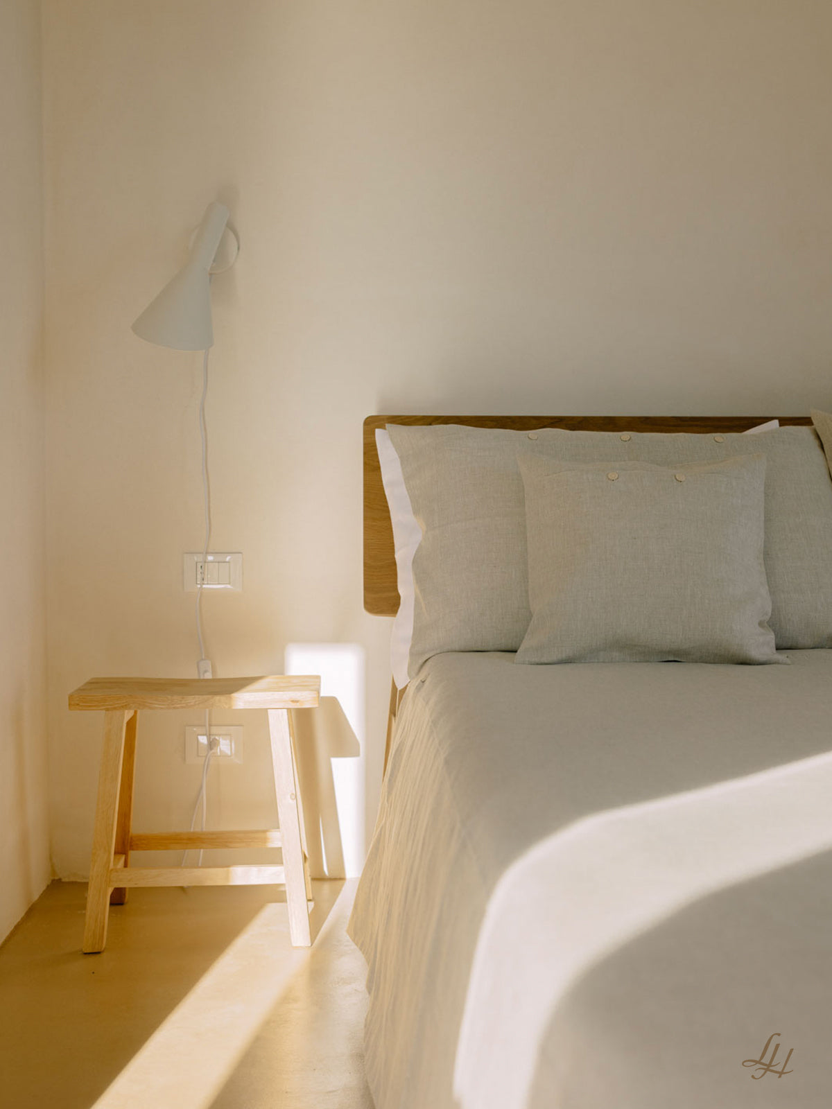 Ein ordentlich gemachtes Bett mit natürlicher Bettwäsche neben einem Holzhocker und einer weißen Wandlampe in einem sonnendurchfluteten, minimalistischen Raum.