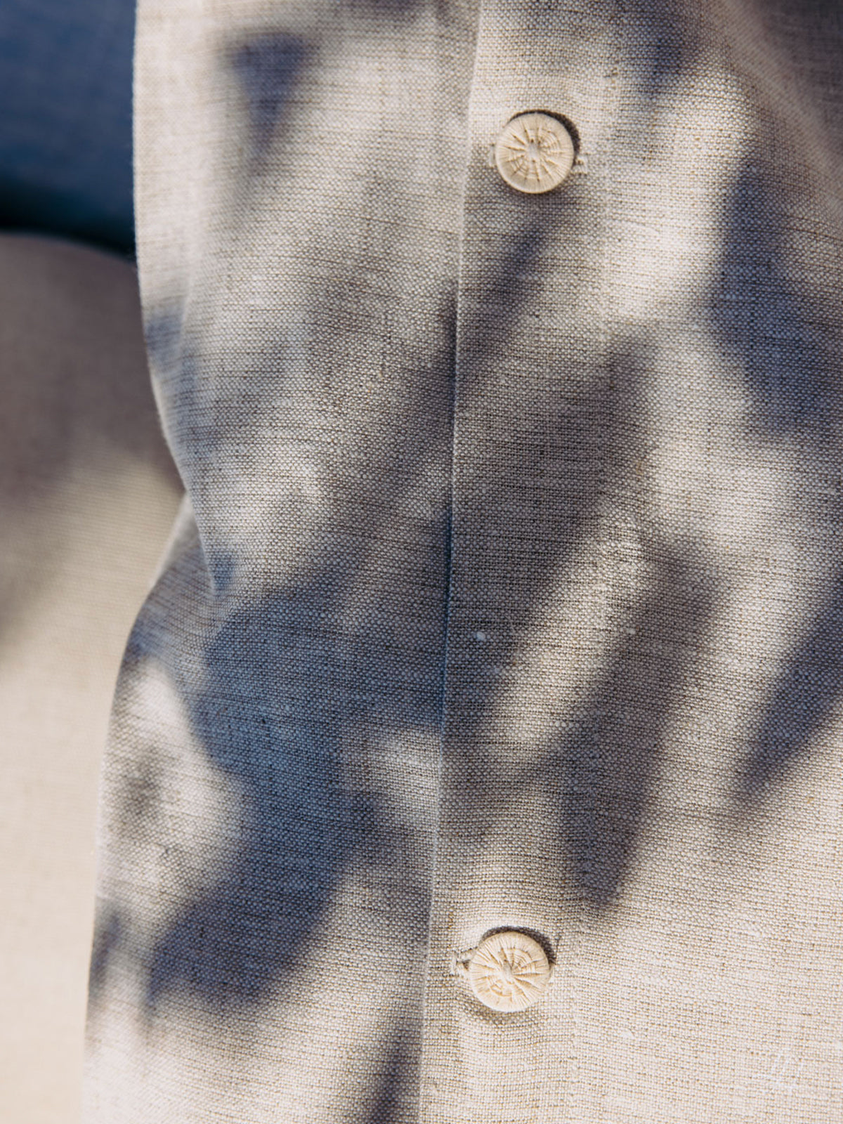 Nahaufnahme eines beigen Leinenbettwäsche-Hemdes mit zwei Knöpfen, das durch das nahe gelegene Blattwerk sanft beschattet wird.