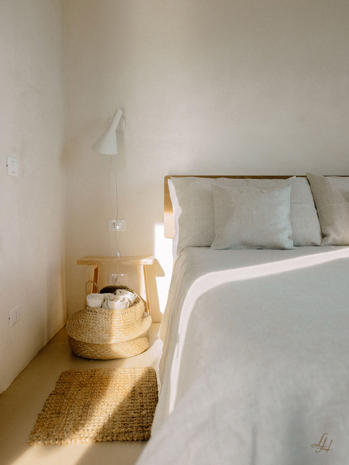 Minimalistisches Schlafzimmer mit einem Bett in Reinleinen-Bettwäsche, weißem Bettzeug, einem Nachttisch aus Holz, einer Lampe, einem Korb und einem kleinen Teppich in natürlichem Licht.