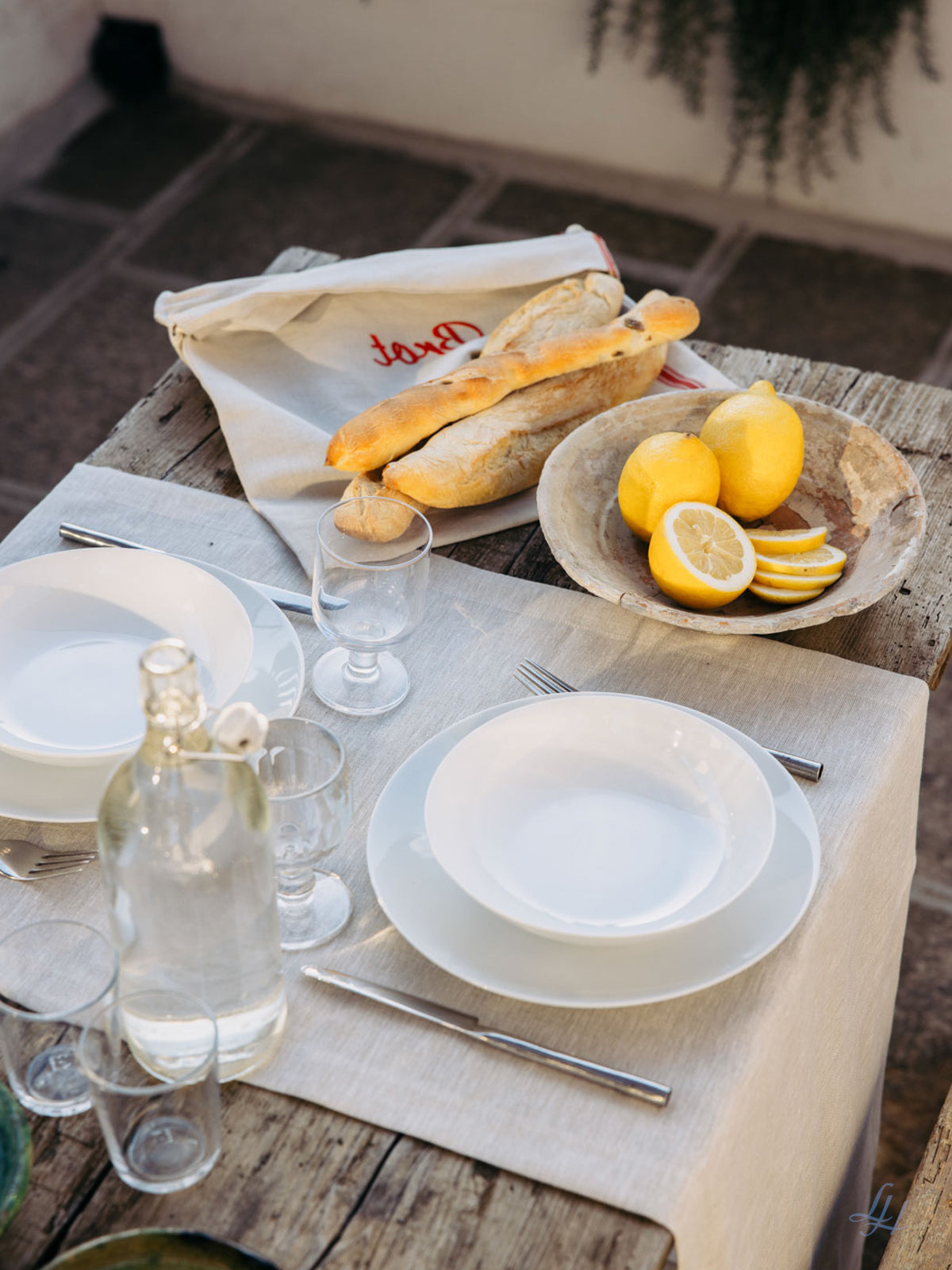 Ein rustikaler Tisch, gedeckt mit Leinen-Tischwäsche, weißen Tellern, Besteck, Wassergläsern, Baguettes und einer Schale mit Zitronen.