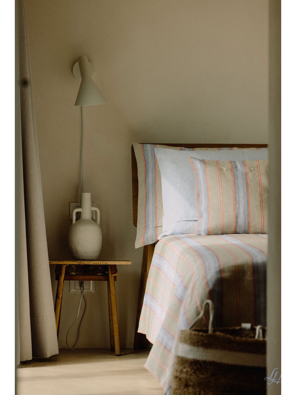 Ein ordentlich gemachtes Bett mit Blockstreifenbettwäsche neben einem hölzernen Nachttisch mit einer weißen Keramikvase und einer Wandlampe.
