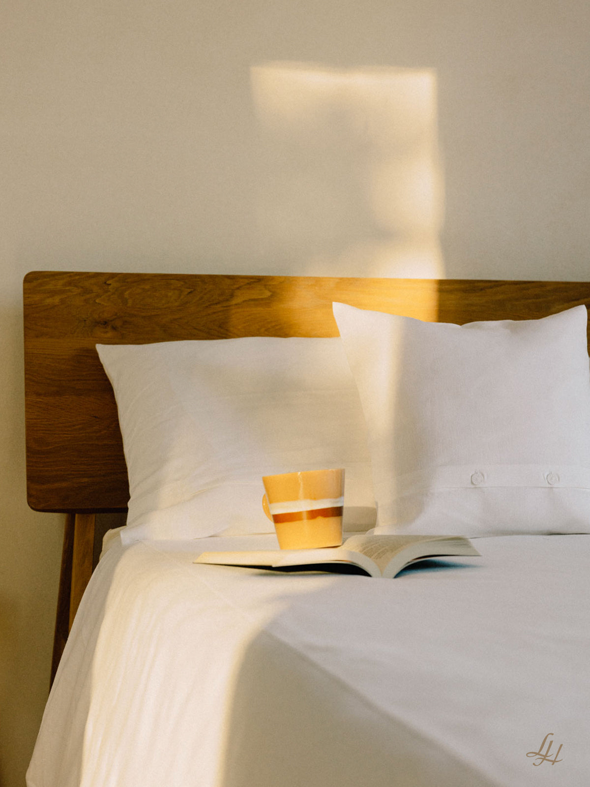Ein gestreifter Becher und ein aufgeschlagenes Buch liegen auf einem Bett mit weißer Bettwäsche, Sonnenlicht wärmt die Wand dahinter.