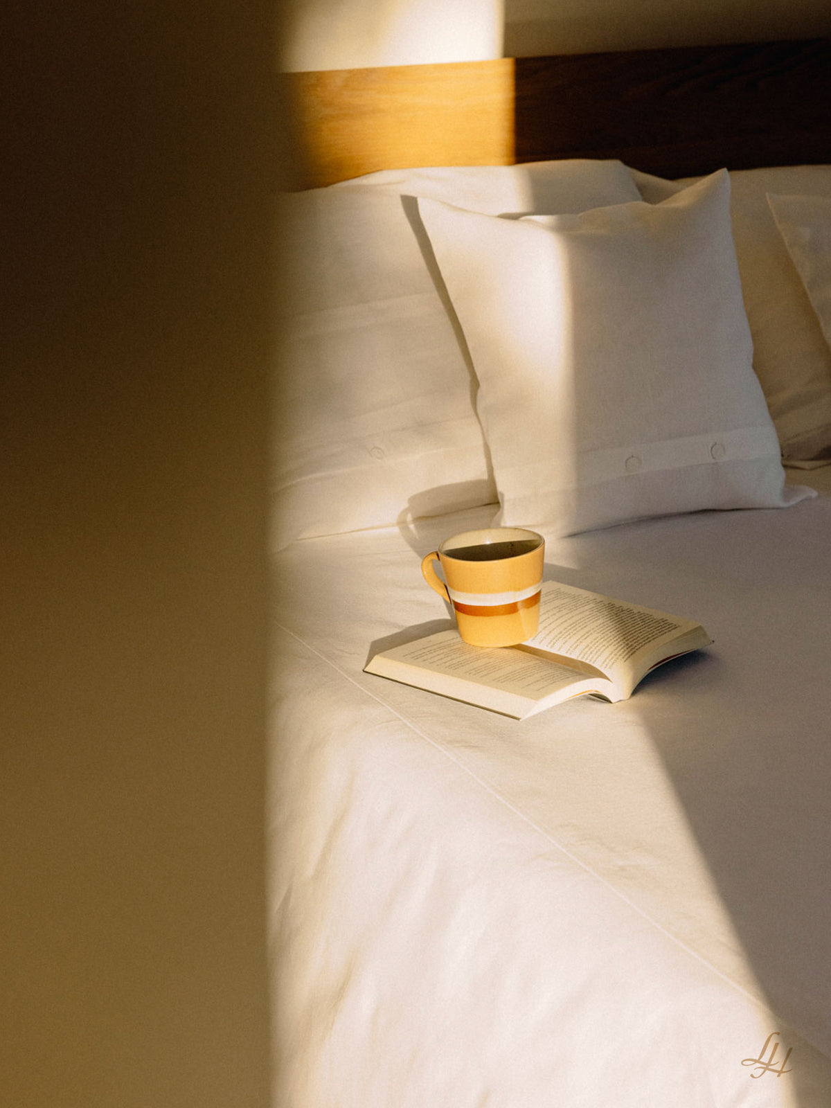 Ein gelb-weißer Becher und ein aufgeschlagenes Buch liegen auf einem Bett mit weißer Bettwäsche, beleuchtet von sanftem Sonnenlicht.