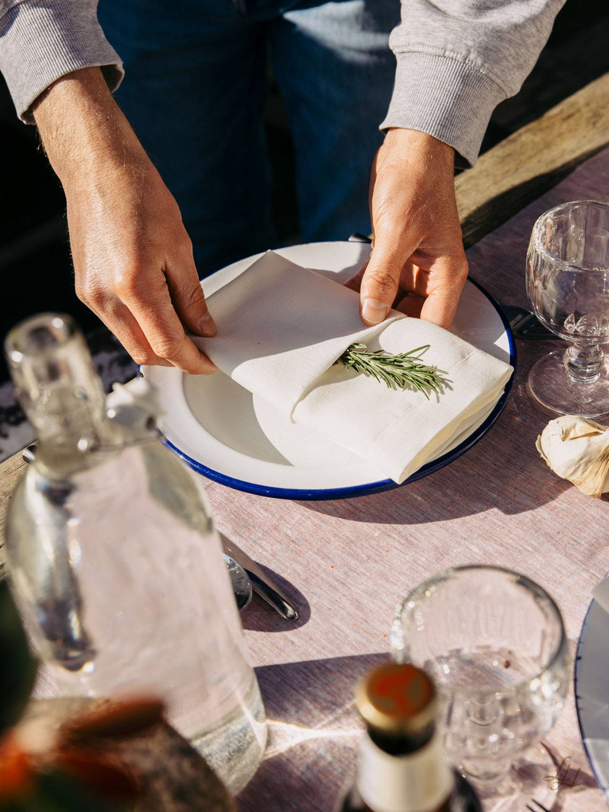 Eine Person faltet eine Leinen Serviette, nachhaltig und hypoallergen, mit Rosmarin auf einem weißen Teller mit Glaswaren und Flasche.