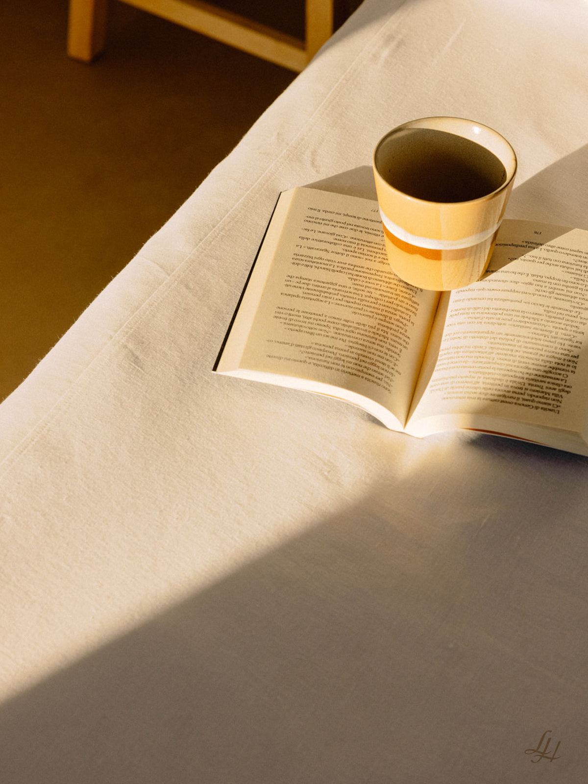 Ein aufgeschlagenes Buch und eine gelb gestreifte Tasse ruhen auf sonnenbeschienener europäischer Flachs weißer Bettwäsche neben einem Holzstuhl.