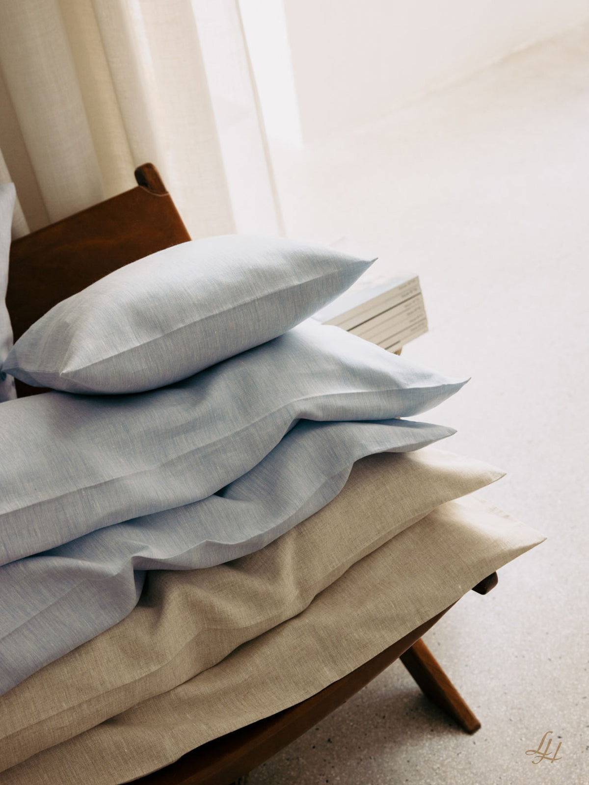 Ein Stapel hellblauer und beiger Kissen aus Leinen Bettwäsche liegt auf einem braunen Stuhl neben weißen Büchern und durchsichtigen Vorhängen.