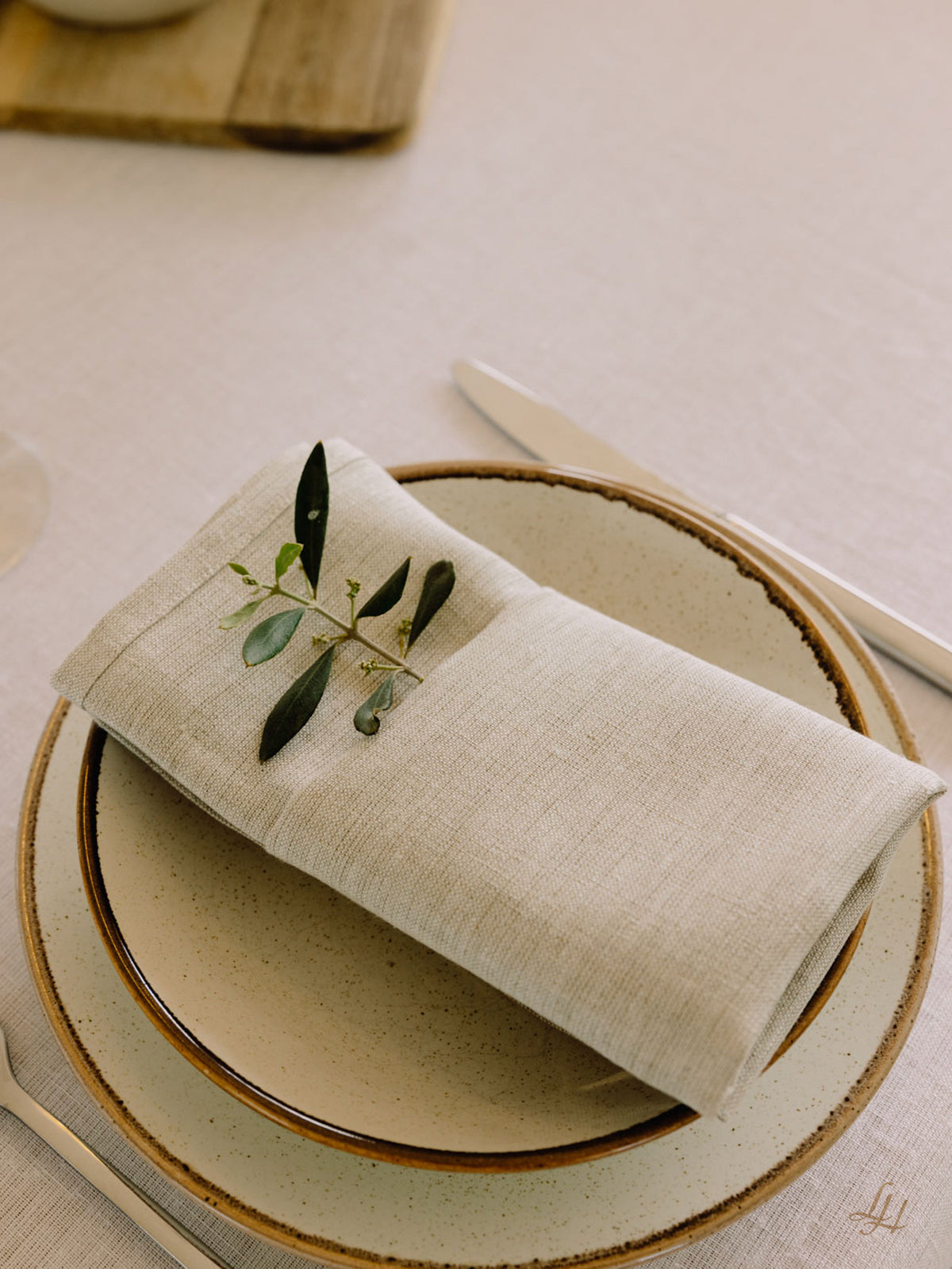 Eine gefaltete beigefarbene Leinenserviette mit grünen Blättern liegt auf gestapelten neutralen Tellern auf einem hellen Tischtuch, daneben liegt das Silberbesteck.