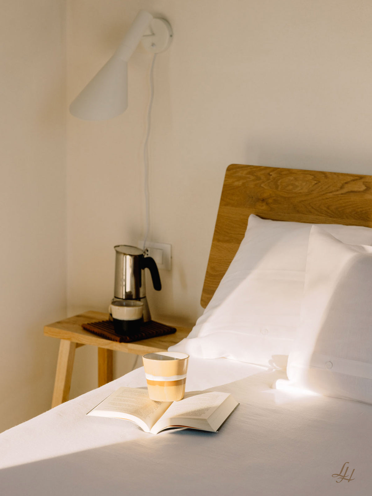Eine Tasse und ein aufgeschlagenes Buch stehen auf einem Bett mit weißer Bettwäsche neben einem hölzernen Nachttisch mit einer Kaffeemaschine und einer Lampe darüber.