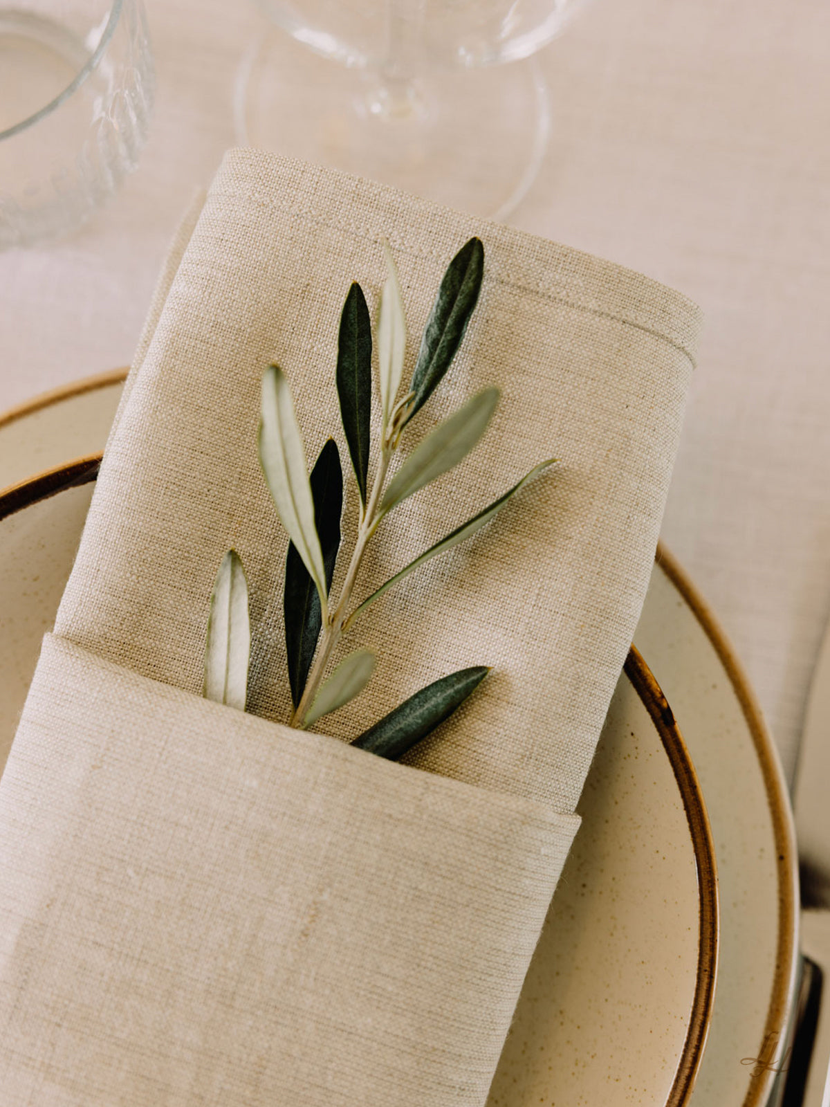 Eine beigefarbene Leinenserviette mit einem Zweig Olivenblätter wird gefaltet und auf einen cremefarbenen Teller mit braunem Rand gestellt.