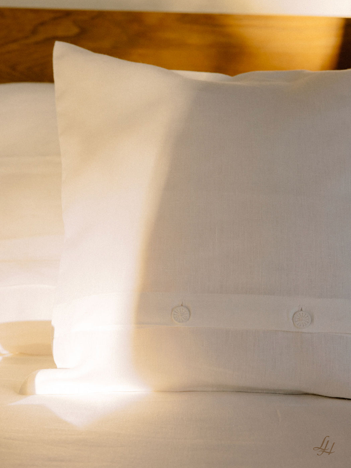 Weiße Bettwäsche aus europäischem Flachs schmückt das Bett, Sonnenlicht spielt über Kissen und hölzernes Kopfteil.