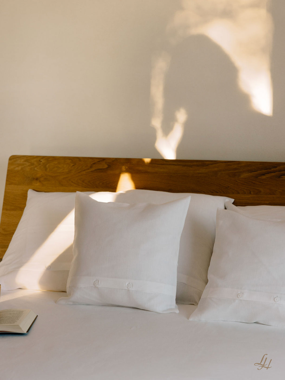 Ein gemachtes Bett mit drei weißen Kissen, europäischer Flachs Leinen Bettwäsche, einem offenen Buch und einem Schatten an der Wand.