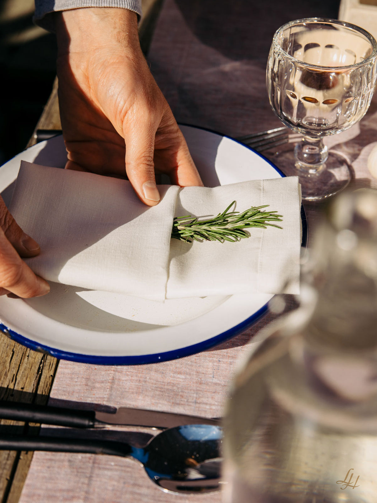 Eine Person stellt eine Leinenmanufaktur Halbleinen Serviette mit Rosmarin auf einen Teller neben Glas und Besteck auf einen gedeckten Tisch.