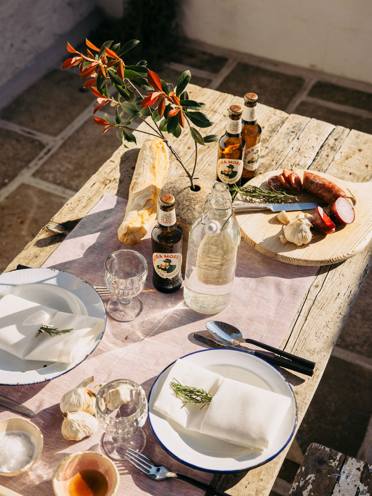 Ein rustikaler Tisch im Freien, gedeckt mit Leinen-Tischwäsche, Tellern, Bierflaschen, Brot, Wurst, Knoblauch und einem Zweig Rosmarin.