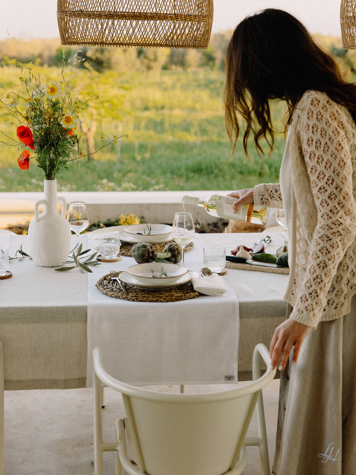 Eine Frau gießt ein Getränk an einem Tisch im Freien ein, der mit einem Halbleinen-Tischläufer, Tellern, Gläsern, Blumen und Artischocken gedeckt ist.