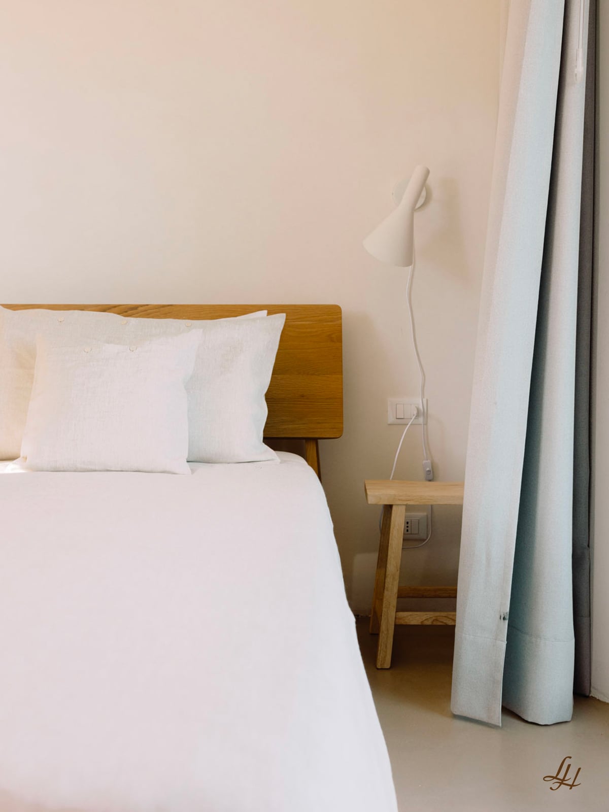 Ein ordentlich gemachtes Bett mit atmungsaktiv weißer Bettwäsche, einem hölzernen Kopfteil, Beistelltisch, weißer Lampe und hellen Vorhängen.