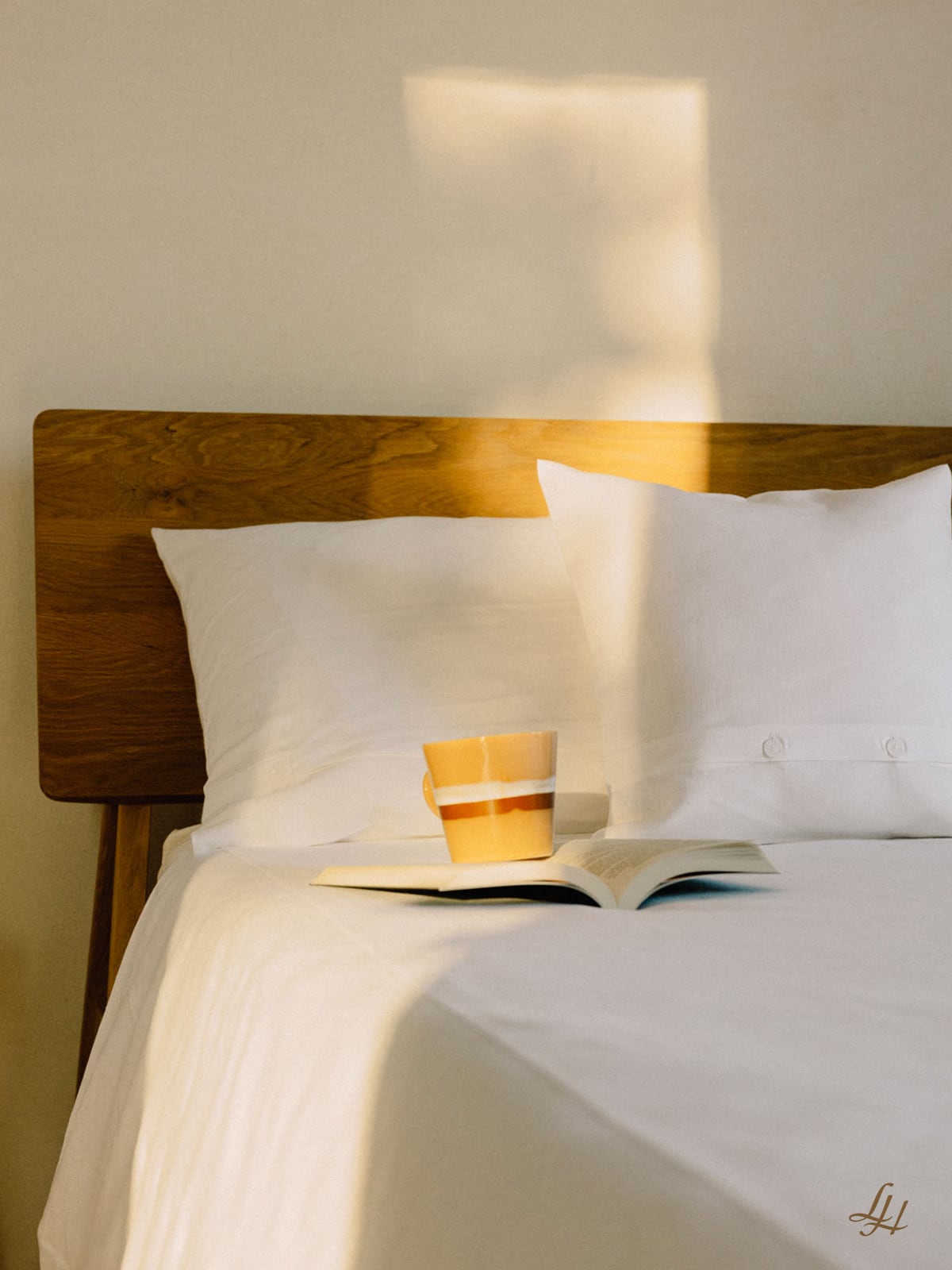 Eine Keramiktasse und ein aufgeschlagenes Buch sitzen auf weißem Leinen mit Kissenbezug, getaucht in weiches, atmungsaktives Tageslicht.