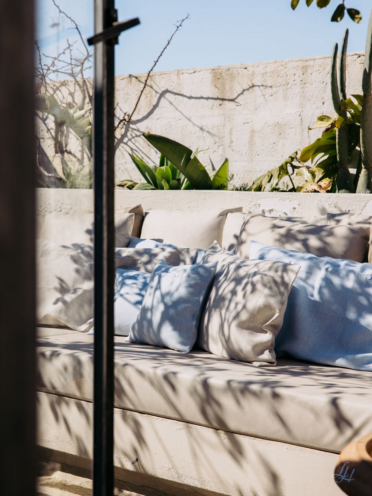 Beigefarbenes Outdoor-Sofa mit hypoallergenen Leinen-Kissenbezügen, umgeben von Pflanzen und Sonnenlicht, das Schatten auf die Kissen wirft.