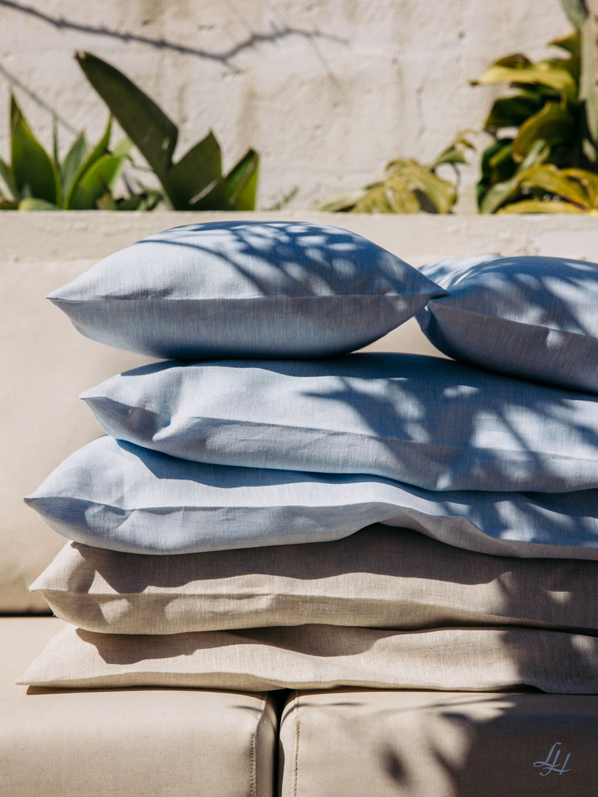Vier gestapelte Kissen in Hellblau und Beige mit Leinen-Kissenbezug auf einem Outdoor-Sofa, das von Blätterschatten beschattet wird.
