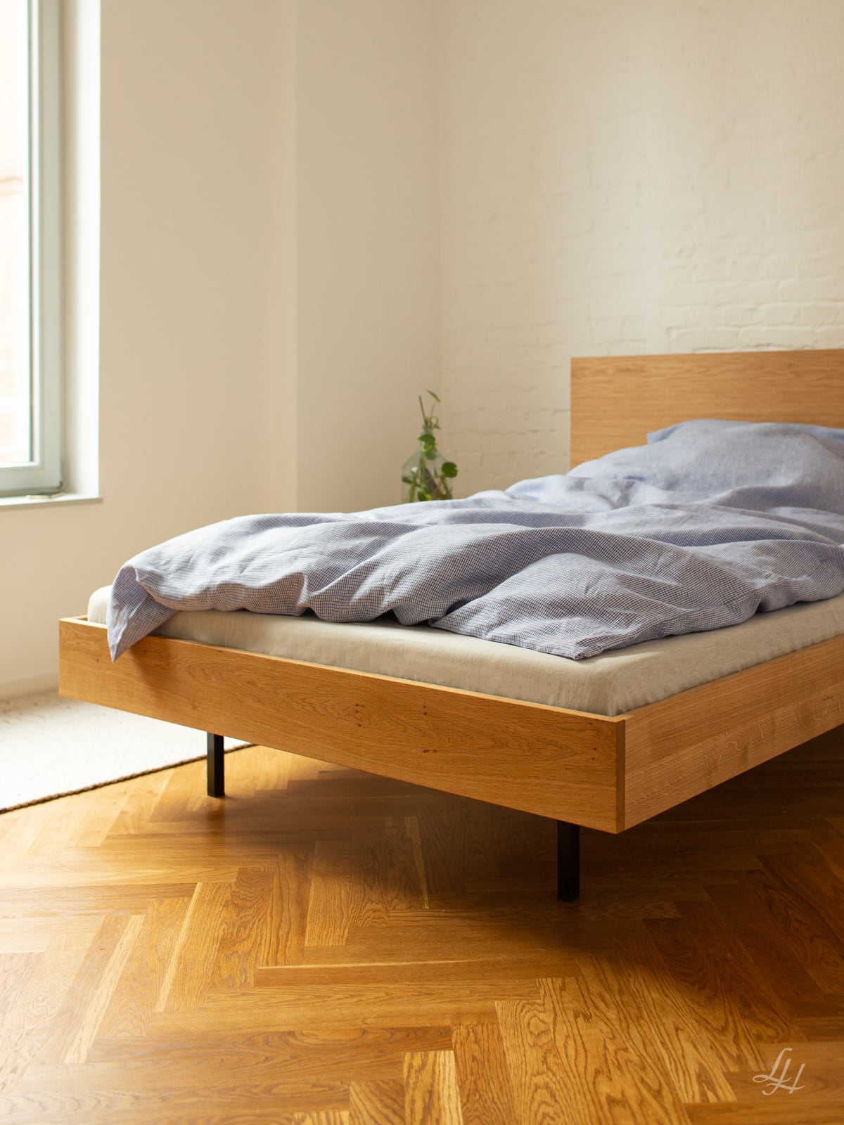 Ein modernes Holzbett mit blauem Bauernkaro Bettwäsche-Set steht in einem hellen Raum mit Holzboden und einer kleinen Pflanze an der Wand. Foto von: © Sebastian Fengler