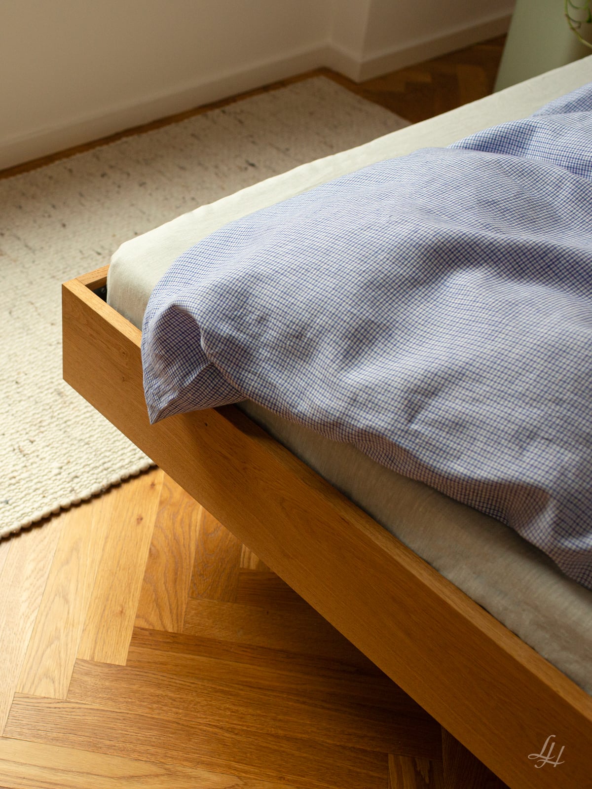 Ein hölzerner Bettrahmen mit blau-weißer Bauernkaro-Bettwäsche neben einem gewebten Teppich auf einem Parkettboden, perfekt für den Landhausstil-Charme. Foto von: © Sebastian Fengler