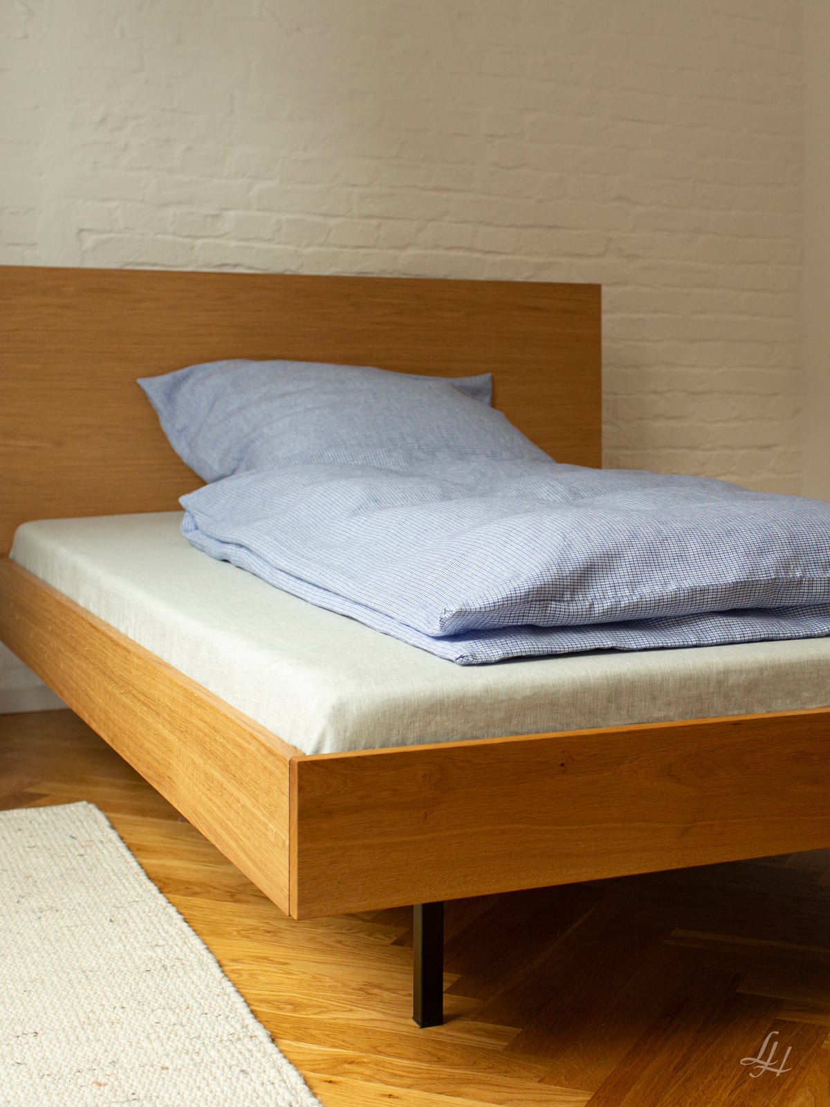 Ein Holzbett mit hellblauem Leinen Bettwäsche-Set in einem minimalistischen Zimmer mit weißer Ziegelwand und Holzboden. Foto von: © Sebastian Fengler