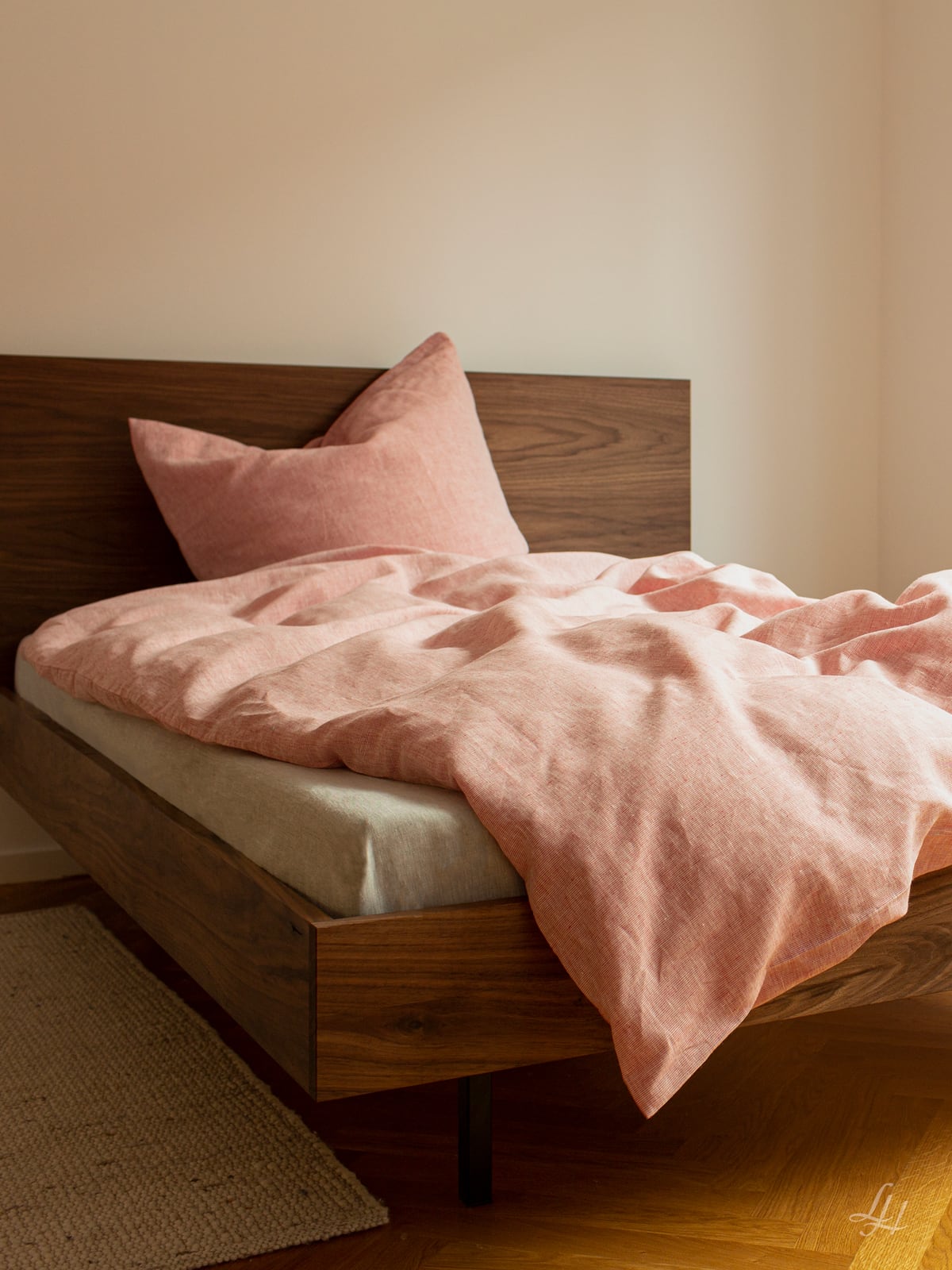 Ein Holzbett mit rosa Leinenbettwäsche und einem Kissen steht in einem einfachen Zimmer mit einem beigen Teppich und hellen Wänden. Foto von: © Sebastian Fengler