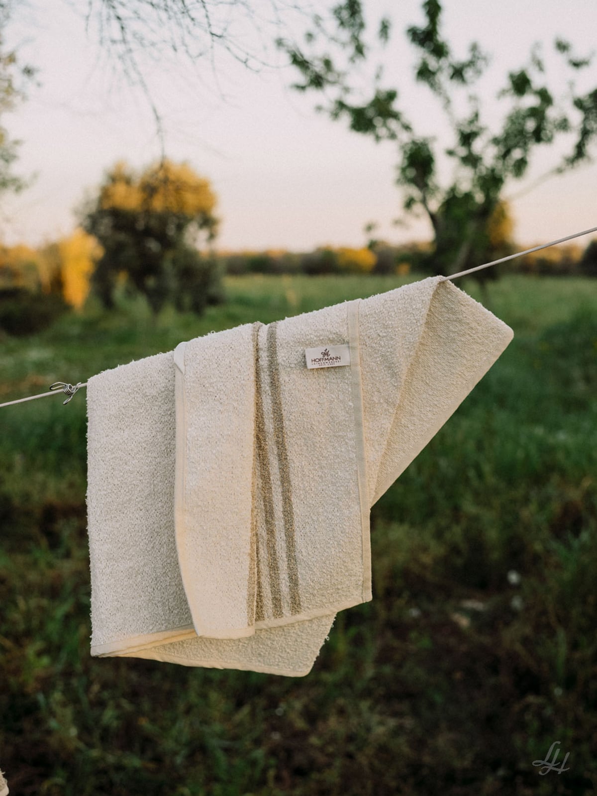 Zwei beigefarbene Leinenfrotté-Saunatücher mit Streifenmuster hängen an einer Wäscheleine im Freien, im Hintergrund Bäume und Gras.