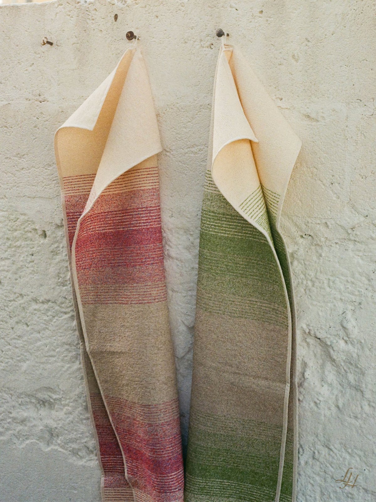 Zwei gewebte Textilien, darunter ein Leinenhandtuch mit roten Streifen, hängen nebeneinander an einer hell strukturierten Wand.