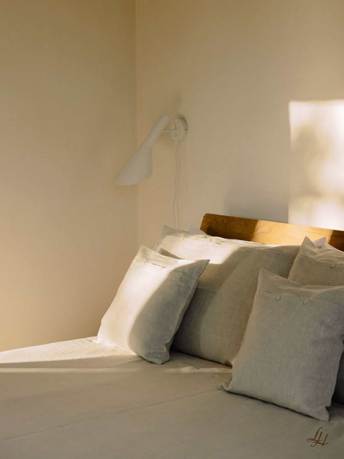 Ein ordentlich gemachtes Bett mit Leinen-Kissenbezug und beigen Kissen steht vor einer cremefarbenen Wand unter einer weißen Wandlampe.