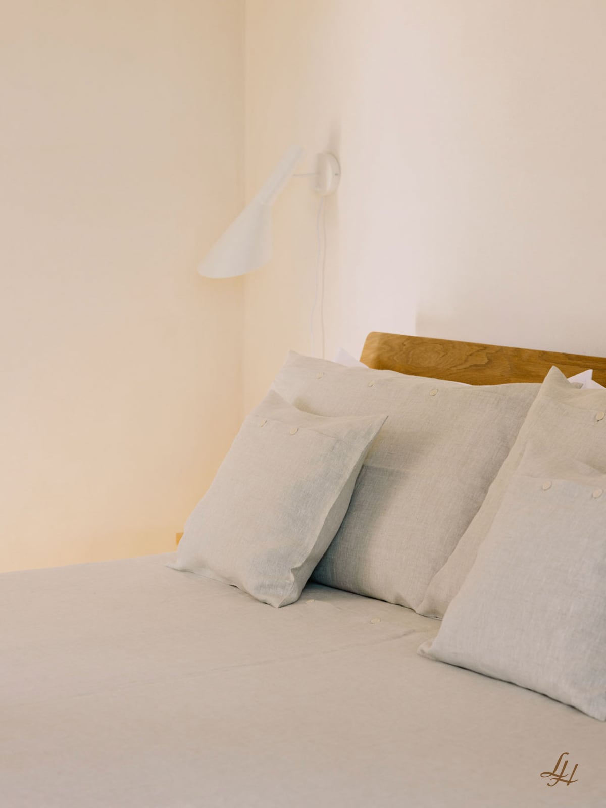 Ein ordentlich gemachtes Bett mit Naturtönen, hellgrauem Leinen-Kissenbezug, einem hölzernen Kopfteil und einer weißen Wandlampe darüber.