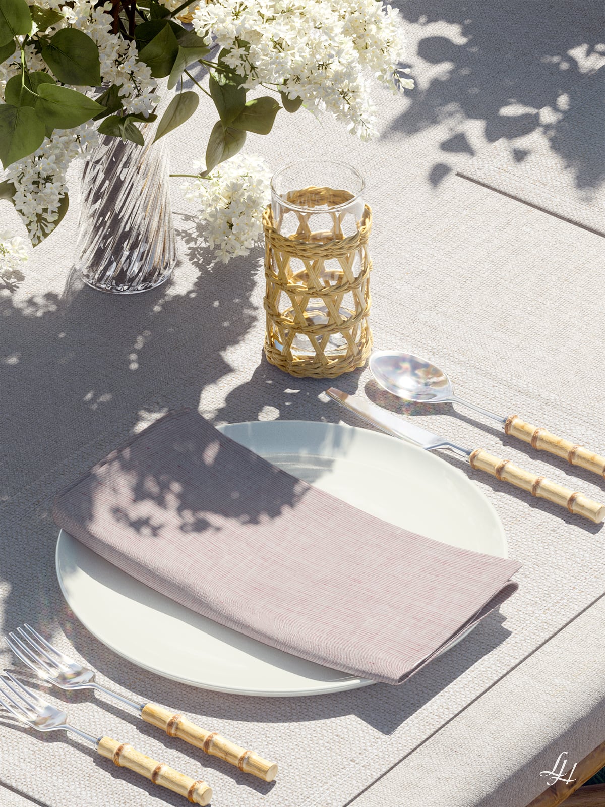 Ein gedeckter Tisch mit Leinenservietten, Goldbesteck, einem Glas mit geflochtenem Halter und einer Vase mit weißen Blumen.