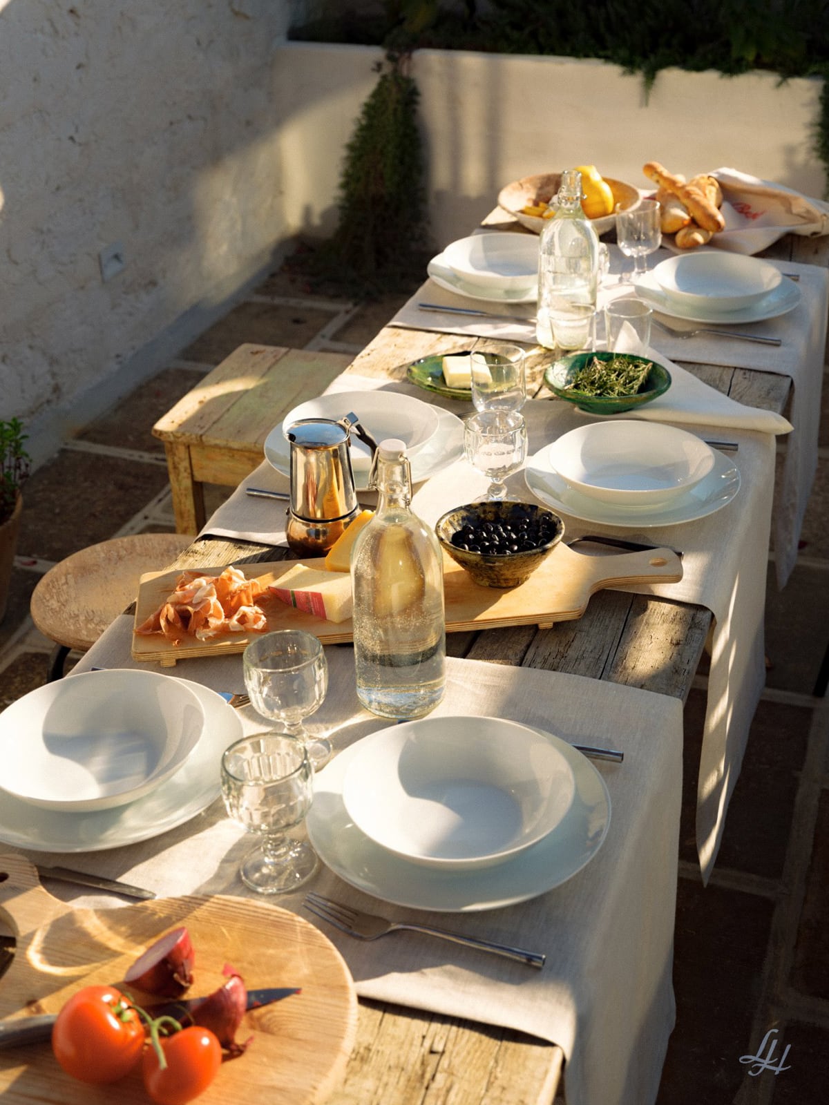 Für eine Mahlzeit gedeckter Tisch im Freien, geschmückt mit Leinen-Tischwäsche, Tellern, Gläsern, Wasserflaschen, Brot, Fleisch, Oliven und Feigen.