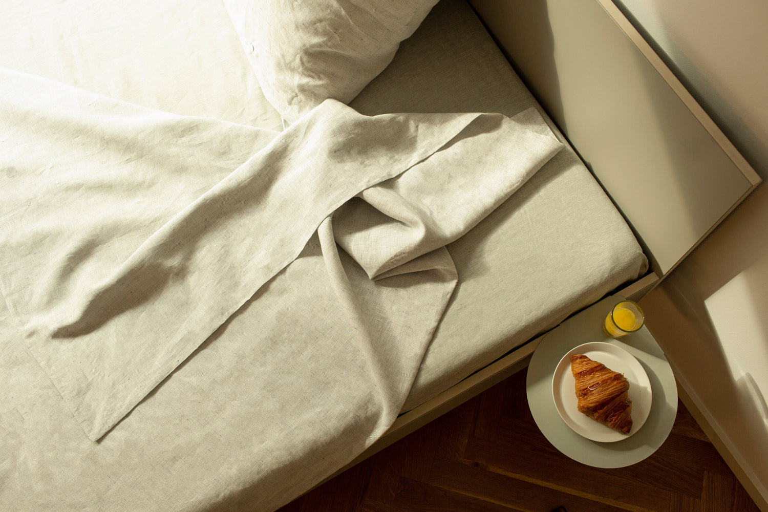 Bett mit Bettwäsche aus Leinen und einem Nachttisch daneben, auf dem Croissant und Saft stehen.