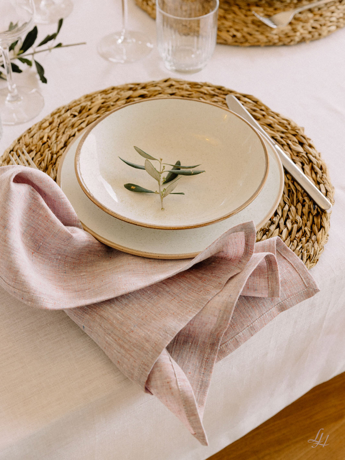 Ein gedeckter Tisch mit einem weißen Teller, einer Schale, Leinen-Servietten, einer rosa Serviette und Grünzeug auf einem nachhaltig gewebten Platzdeckchen.