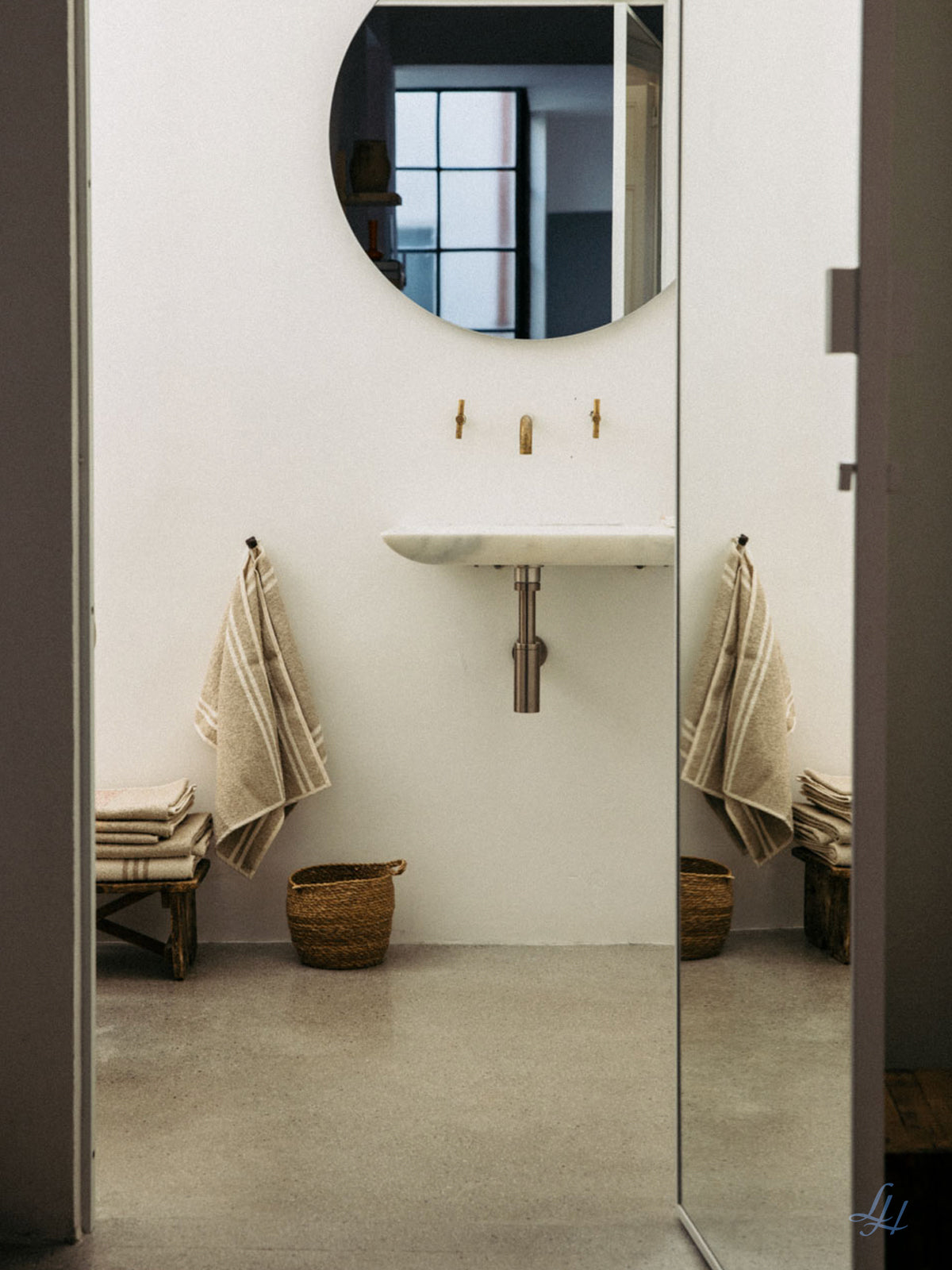 Ein minimalistisches Badezimmer mit einem runden Spiegel, einem Wandwaschbecken und Leinenfrotté-Handtüchern, die auf einer kleinen Bank ordentlich gefaltet sind.