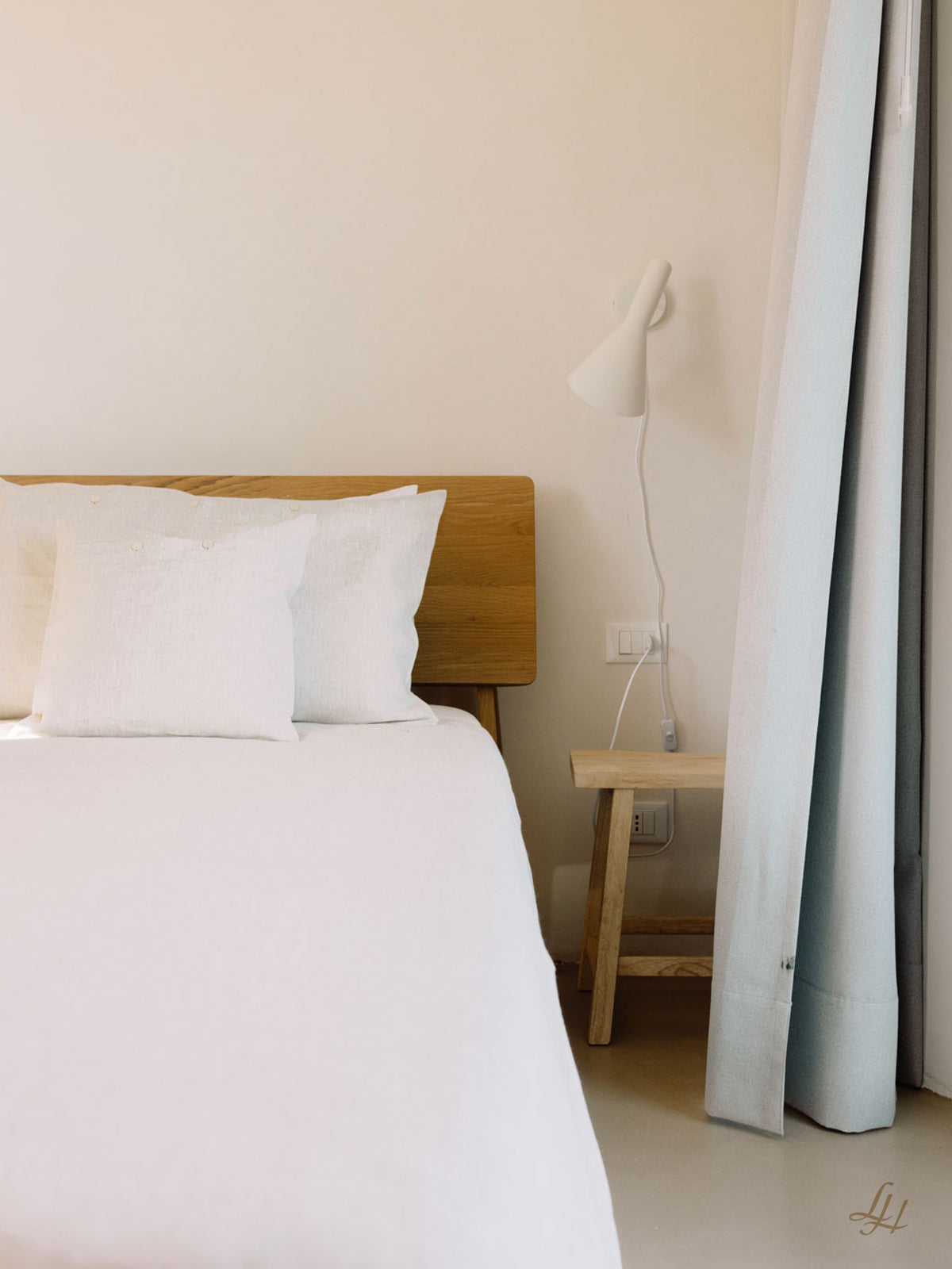 Ein ordentlich gemachtes Bett mit weißer Bettwäsche aus europäischem Flachs, Kopfteil aus Holz, Beistelltisch und weißer Wandlampe.
