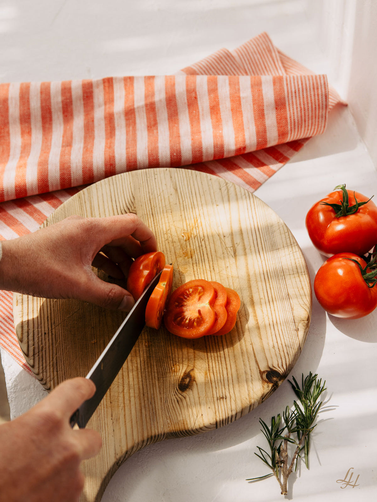 Eine Hand schneidet eine Tomate auf einem Holzbrett neben ganzen Tomaten, Rosmarin und klassischem Streifendesign Leinen Küchentücher.