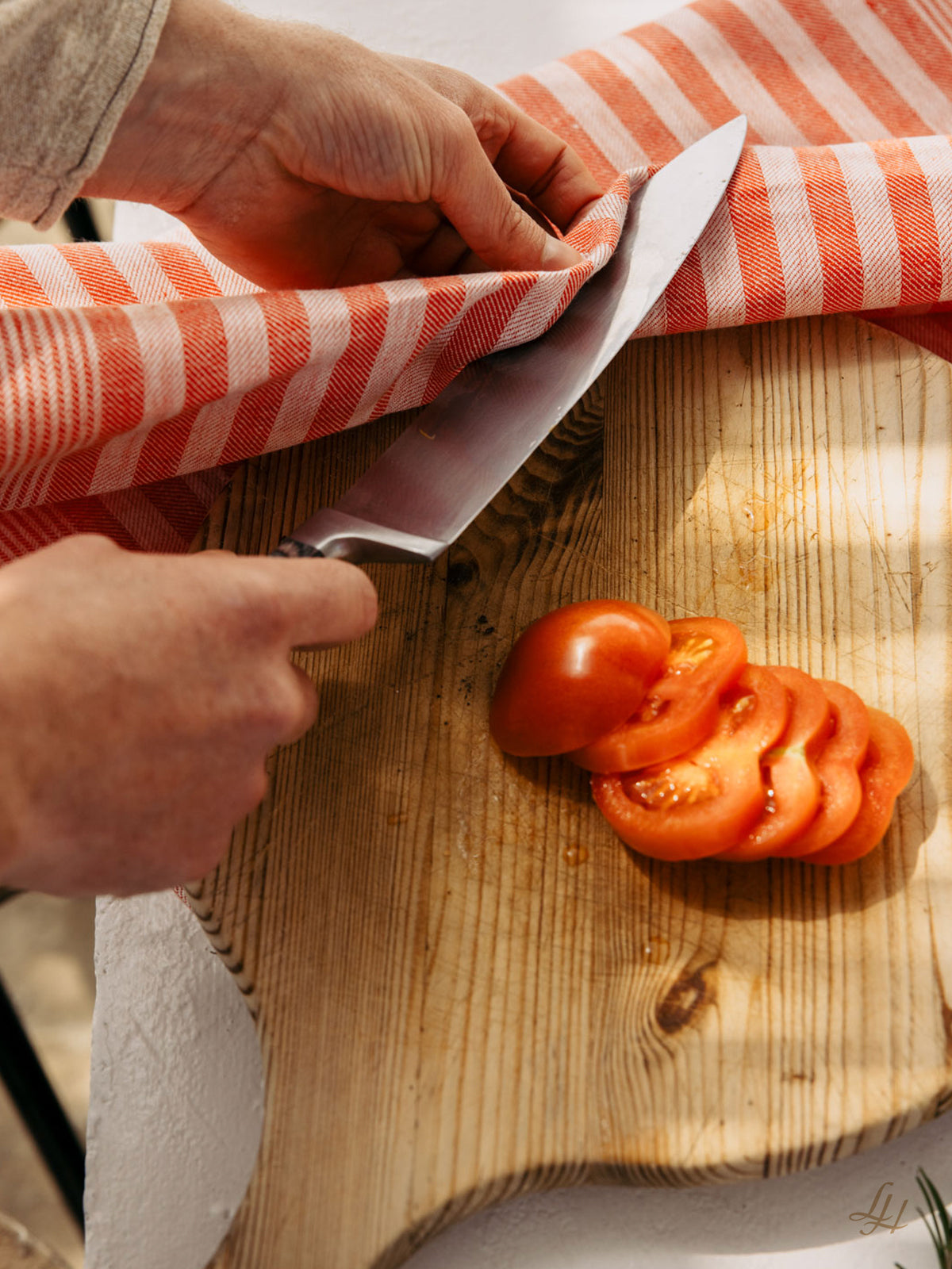 Eine Hand wischt ein Messer mit einem Reinleinen Geschirrtuch in klassischem Streifendesign neben einer geschnittenen Tomate auf einem Holzbrett ab.
